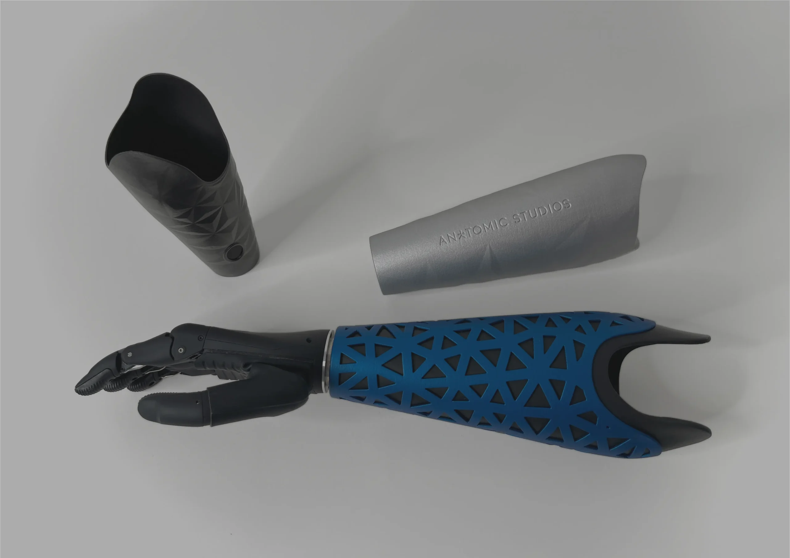 Three prosthetic arm covers by Anatomic Studios, shown alongside a black prosthetic hand. The covers feature different styles, including a black geometric design, a sleek silver version with branding, and a blue mesh-patterned cover attached to the prosthesis.